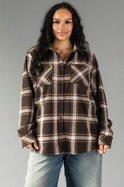 A woman with wavy dark hair wears the Panache Apparel Collection Dark Brown, Cream & Black Plaid Button Up with chest pockets and blue jeans, standing against a plain gray background.