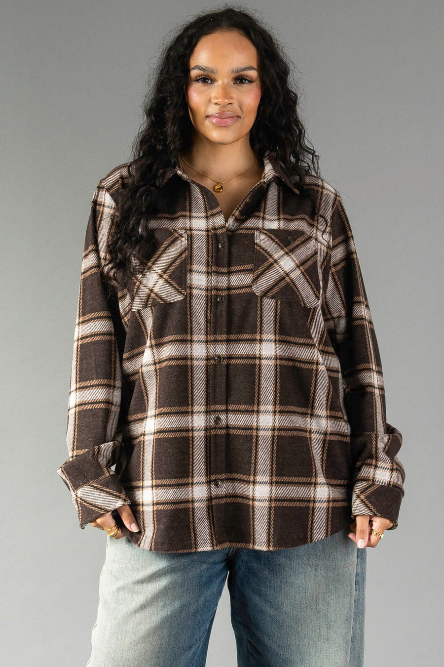 A woman with wavy dark hair wears the Panache Apparel Collection Dark Brown, Cream & Black Plaid Button Up with chest pockets and blue jeans, standing against a plain gray background.