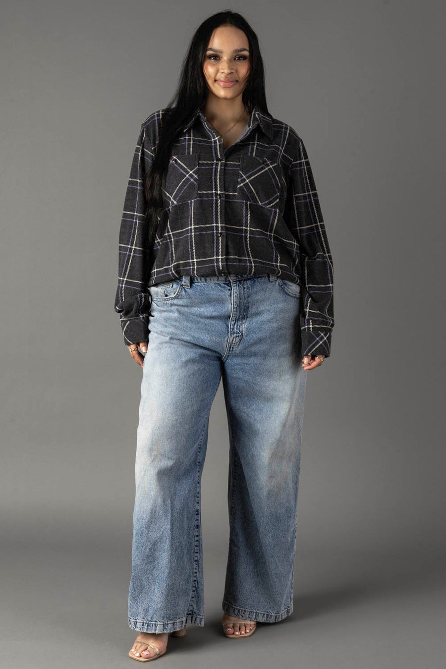 A woman with long dark hair wears the Panache Apparel Collection Dark Gray, Light Blue & White Plaid Button Up and loose, light blue jeans. She stands facing forward, smiling slightly against a plain gray background.