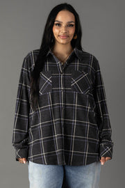 A woman with long dark hair wears the Panache Apparel Collection Dark Gray, Light Blue & White Plaid Button Up and light blue jeans, standing against a plain gray background, facing forward and smiling.
