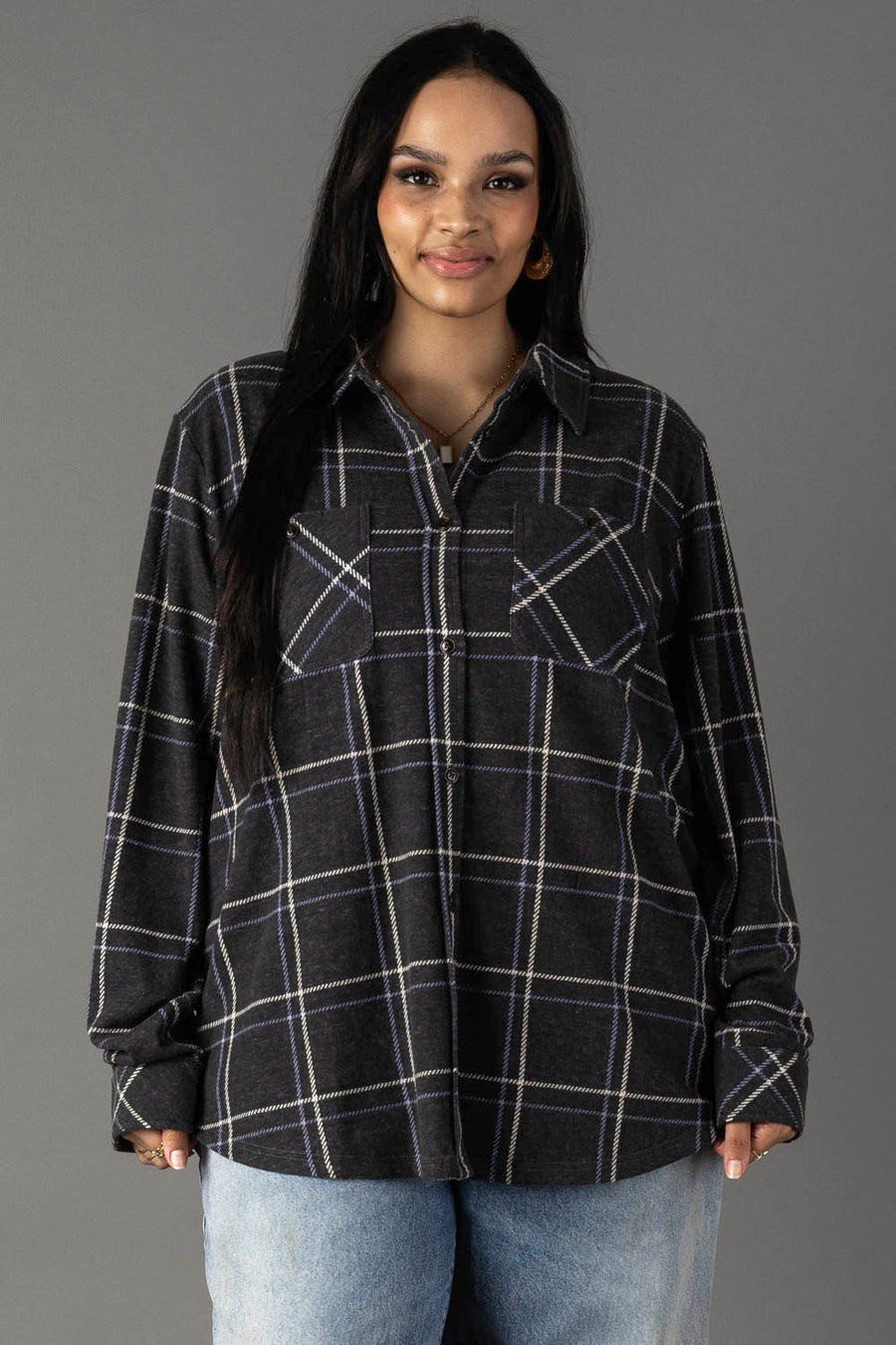 A woman with long dark hair wears the Panache Apparel Collection Dark Gray, Light Blue & White Plaid Button Up and light blue jeans, standing against a plain gray background, facing forward and smiling.