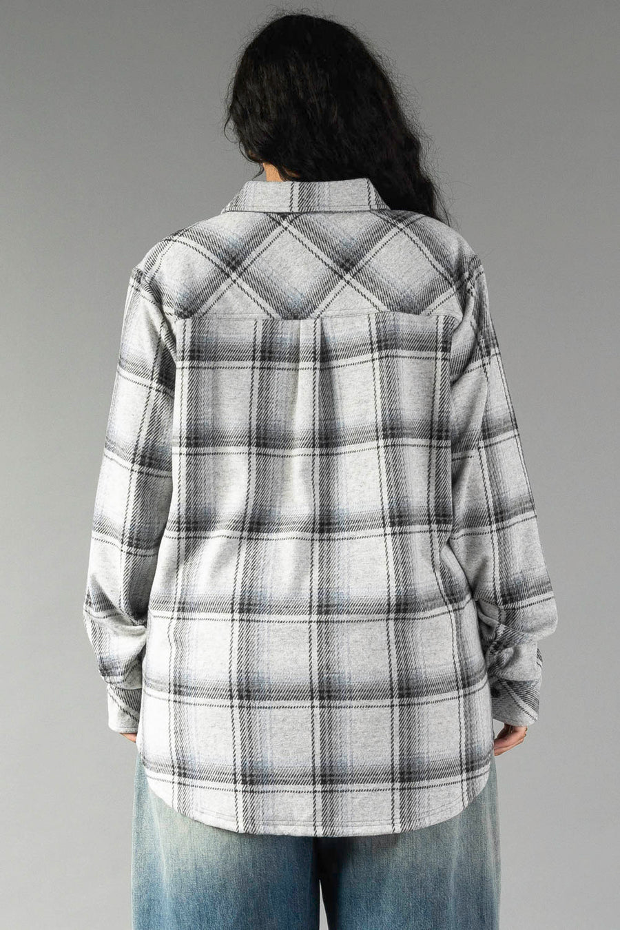 A person with long dark hair is seen from behind, standing against a plain gray background, wearing the Panache Apparel Collection Light Gray, Black & Sage Plaid Button Up along with blue jeans.