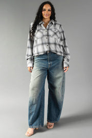 A woman with long dark hair wears the Panache Apparel Collection’s Light Gray, Black & Sage Plaid Button Up and wide-leg blue jeans. She stands against a plain gray background, smiling with her hands relaxed by her sides.