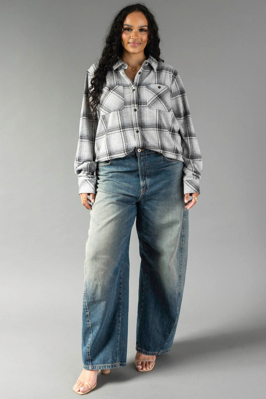 A woman with long dark hair wears the Panache Apparel Collection’s Light Gray, Black & Sage Plaid Button Up and wide-leg blue jeans. She stands against a plain gray background, smiling with her hands relaxed by her sides.