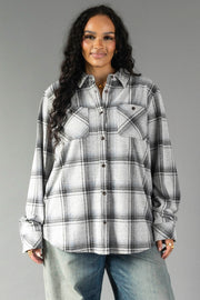 A person with long, wavy dark hair wears the Panache Apparel Collection Light Gray, Black & Sage Plaid Button Up and blue jeans, standing against a plain gray background and smiling slightly at the camera.