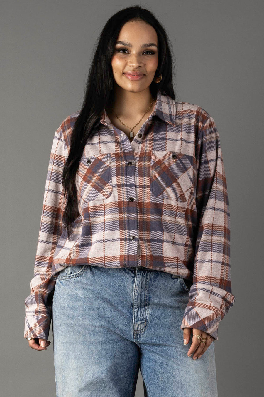A person with long dark hair wears the Panache Apparel Collection’s Dusty Blue, Brick & Tan Plaid Button Up, paired with light blue jeans, standing against a gray background and smiling slightly.