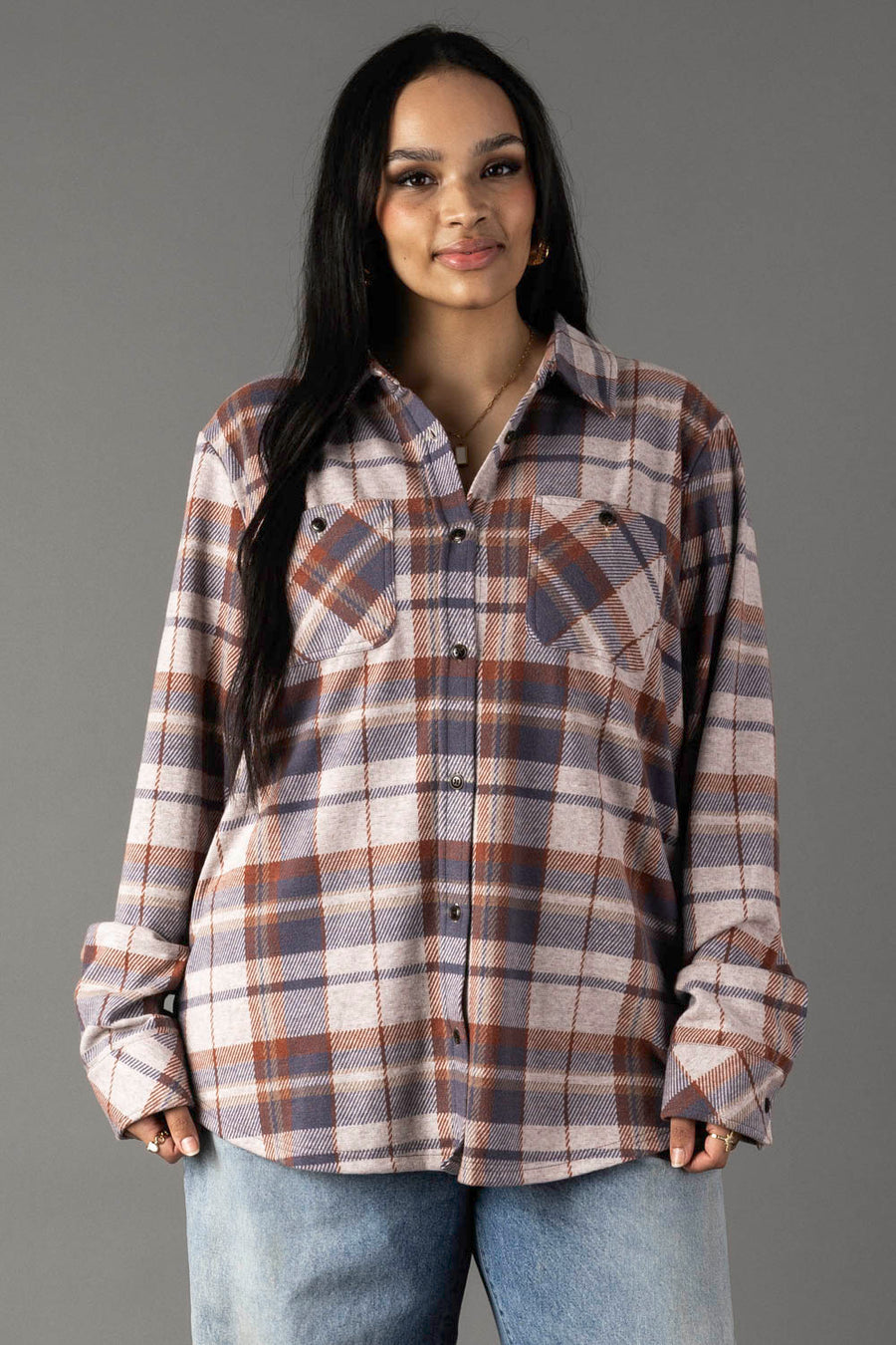 A person with long dark hair wears the Dusty Blue, Brick & Tan Plaid Button Up from Panache Apparel Collection and light blue jeans, standing against a plain gray background and smiling softly at the camera.