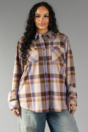 A woman with long curly hair wears the Panache Apparel Collection Dark Brown, Blue & Pink Plaid Button Up with light blue jeans, standing against a plain gray background and smiling slightly.