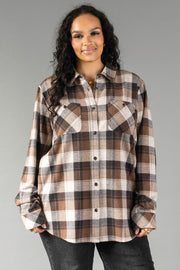 A woman with long curly hair wears the Panache Apparel Collection Brown, Black & Cream Plaid Button Up with dark jeans, standing against a plain gray background and smiling at the camera.