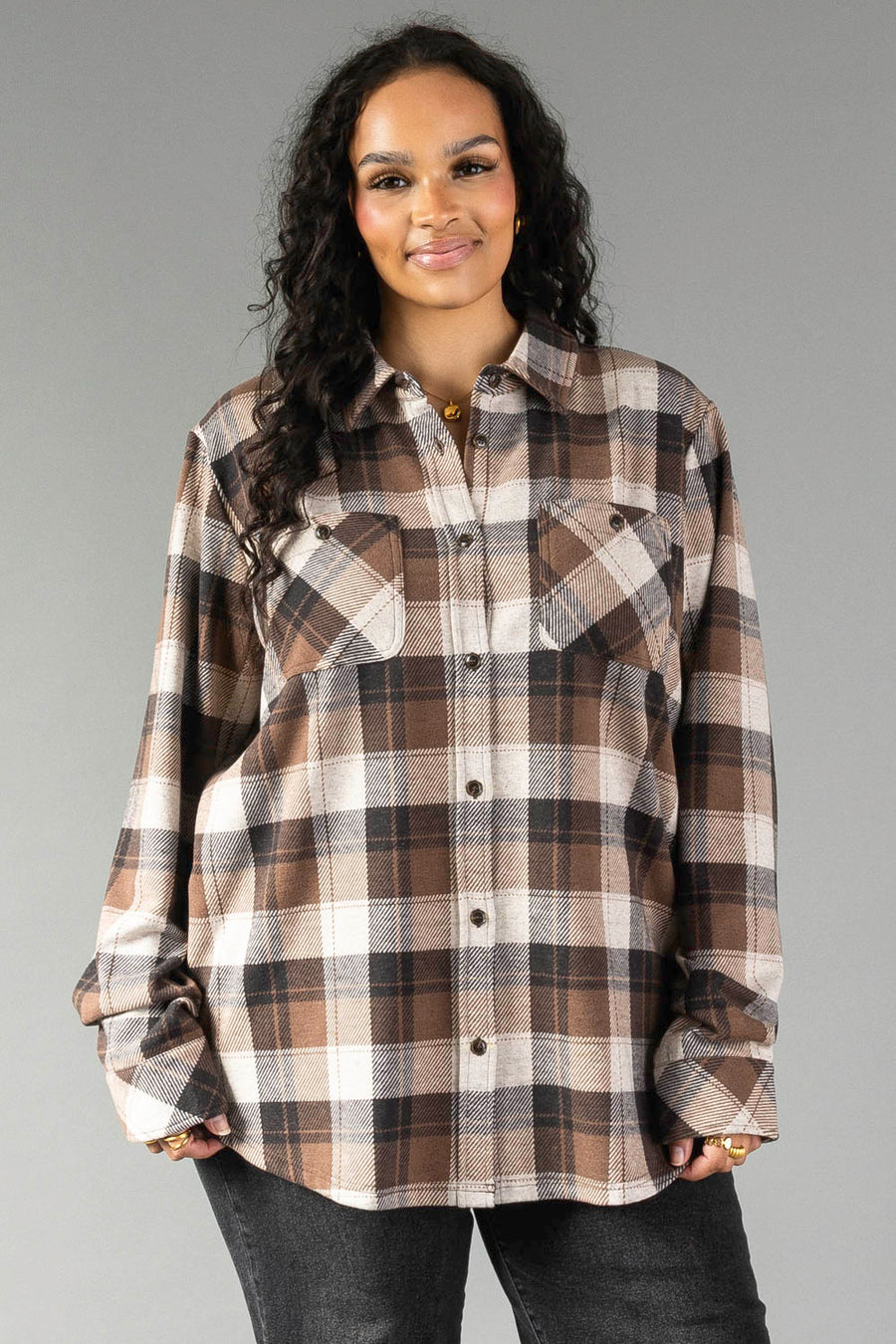 A woman with long curly hair wears the Panache Apparel Collection Brown, Black & Cream Plaid Button Up with dark jeans, standing against a plain gray background and smiling at the camera.