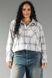 A woman with long curly hair wears the Panache Apparel Collection’s Cream, Dusty Blue & Tan Plaid Button Up paired with high-waisted blue jeans, standing against a plain gray background and smiling gently at the camera.