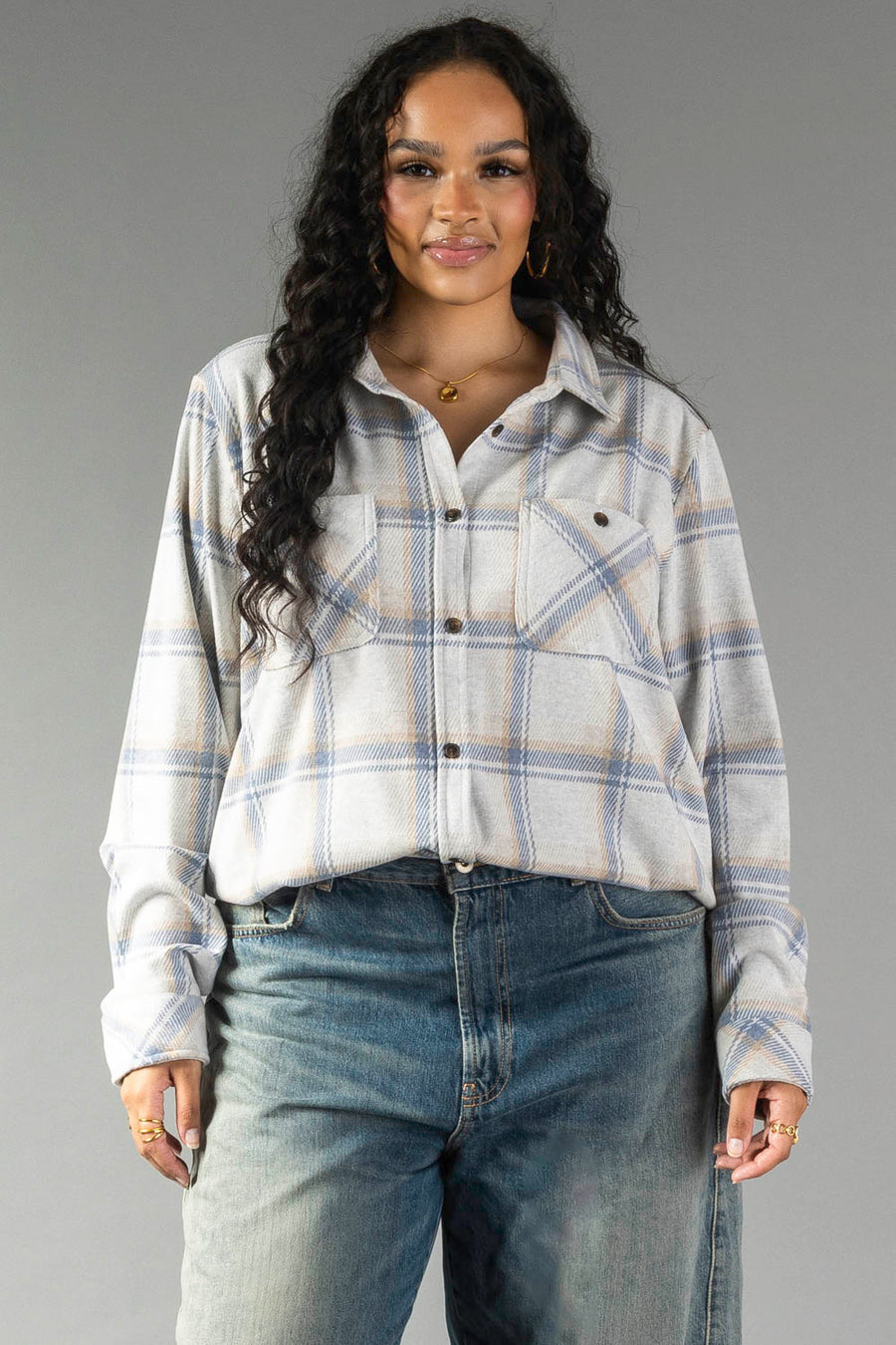 A woman with long curly hair wears the Panache Apparel Collection’s Cream, Dusty Blue & Tan Plaid Button Up paired with high-waisted blue jeans, standing against a plain gray background and smiling gently at the camera.
