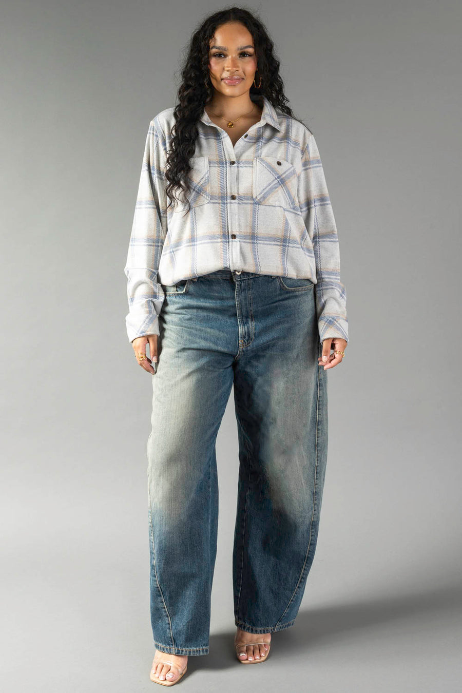 A woman with long dark hair stands against a plain gray background wearing the Panache Apparel Collection’s Cream, Dusty Blue & Tan Plaid Button Up paired with loose-fitting blue jeans. She has a relaxed expression and maintains a neutral stance.