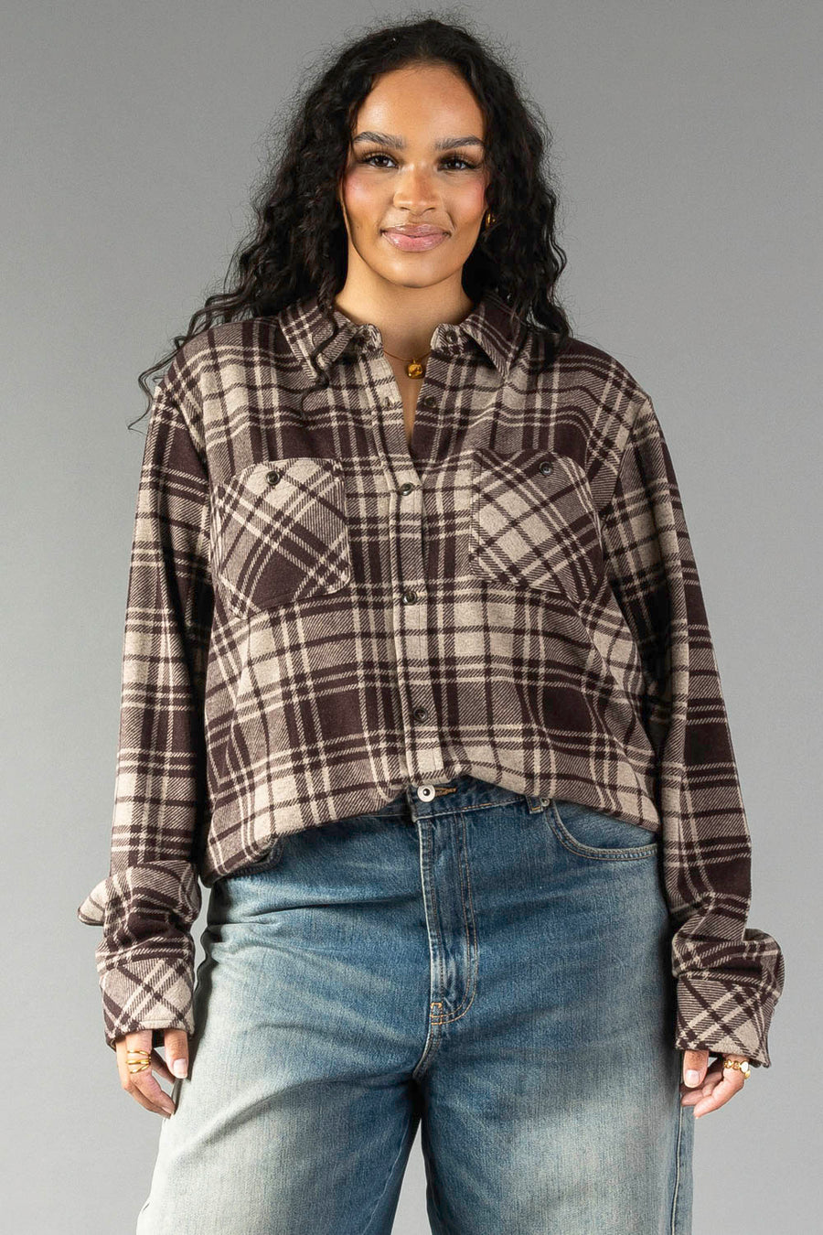 Wearing the Panache Apparel Collection’s Chocolate Brown & Tan Plaid Button Up with blue jeans, a woman with long curly hair stands against a plain gray background, smiling at the camera.