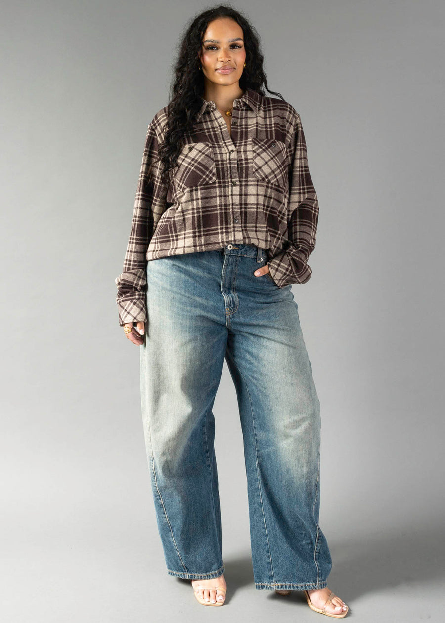 A woman with long dark hair stands facing forward against a plain gray background, wearing the Panache Apparel Collection Chocolate Brown & Tan Plaid Button Up shirt paired with loose blue jeans.