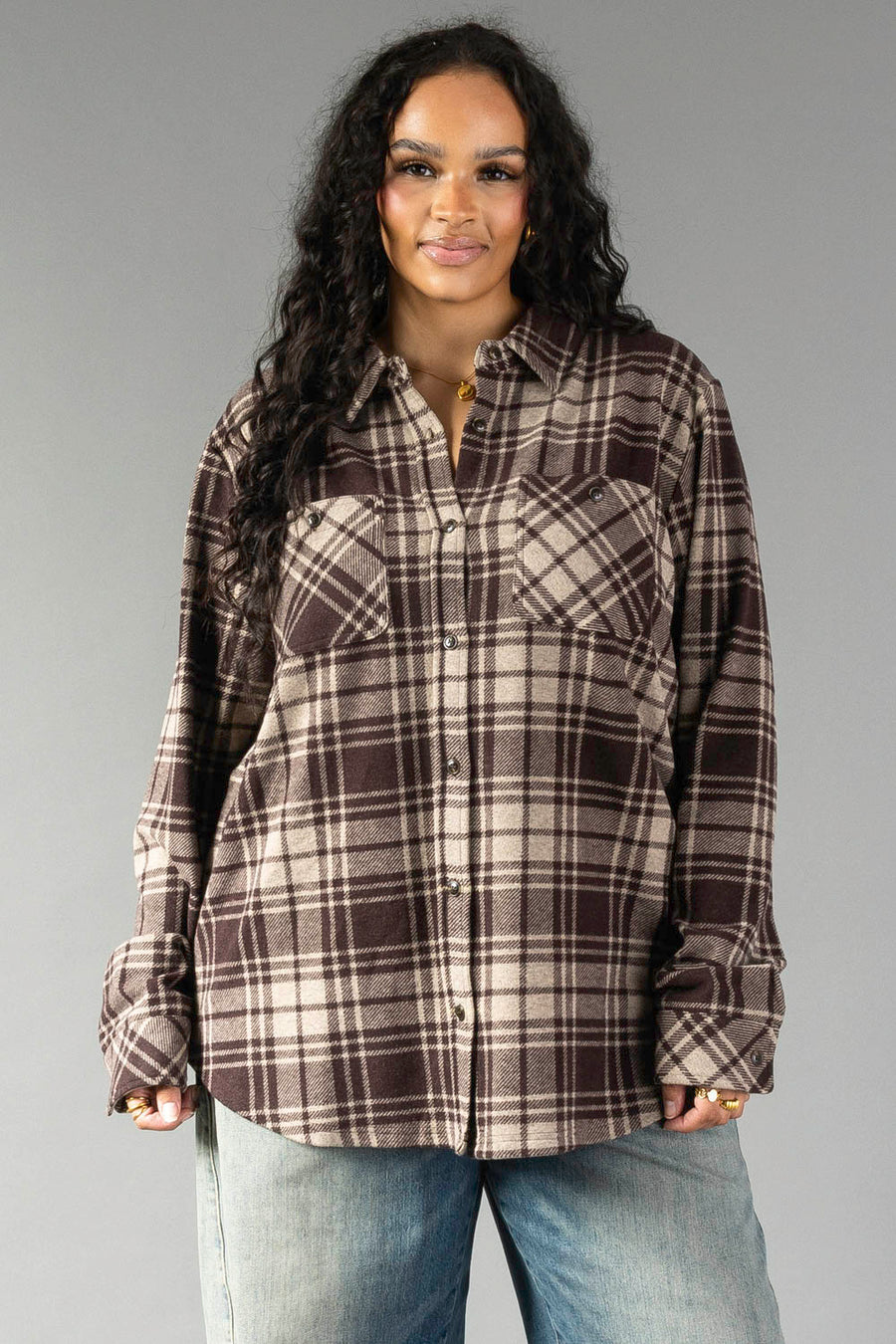 Model is wearing the Panache Apparel Collection’s Chocolate Brown & Tan Plaid Button Up, styled with light blue jeans. She stands against a plain gray background with her long curly hair down, smiling gently at the camera.