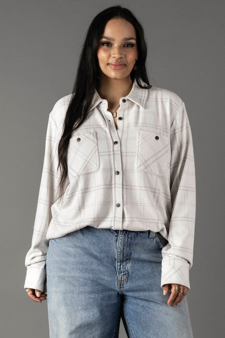 A woman with long dark hair wears the Panache Apparel Collection White & Light Gray Plaid Button Up and light blue jeans, standing and smiling against a plain gray background.