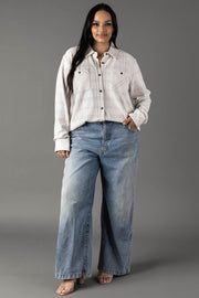 A woman stands against a gray background wearing the Panache Apparel Collection’s White & Light Gray Plaid Button Up paired with loose-fitting, wide-leg blue jeans; one hand in her pocket, she offers a subtle smile.