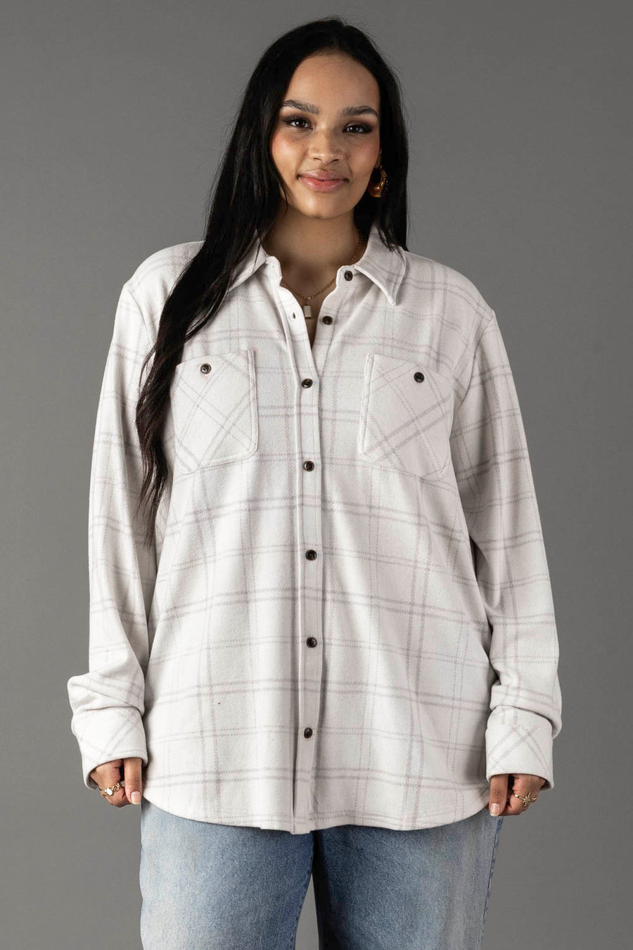 A woman with long dark hair wears the Panache Apparel Collection White & Light Gray Plaid Button Up, featuring front pockets, paired with light blue jeans as she stands against a plain gray background.