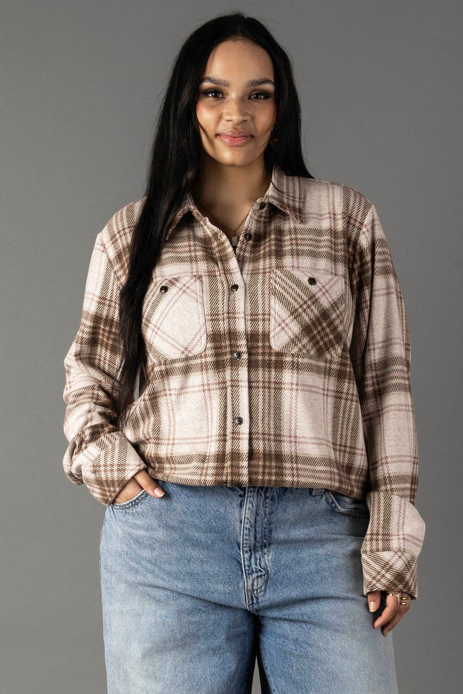 A woman with long dark hair models the Panache Apparel Collection’s Brown, Tan & Rose Plaid Button Up, paired with blue jeans. She stands against a plain gray background and smiles gently at the camera.