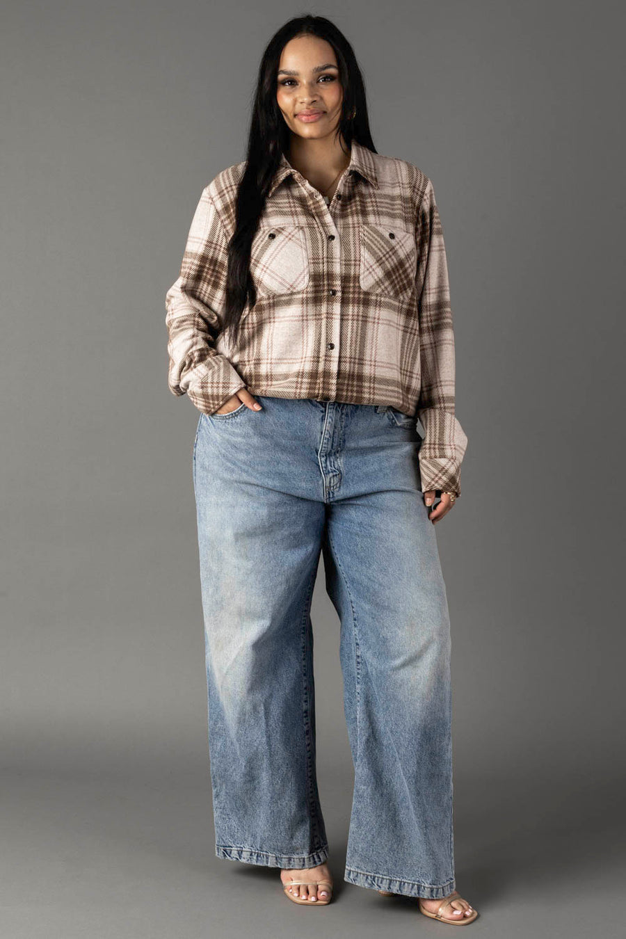 A woman stands against a gray background wearing the Brown, Tan & Rose Plaid Button Up from Panache Apparel Collection, paired with blue wide-leg jeans and sandals. She has long dark hair and is smiling slightly with one hand in her pocket.