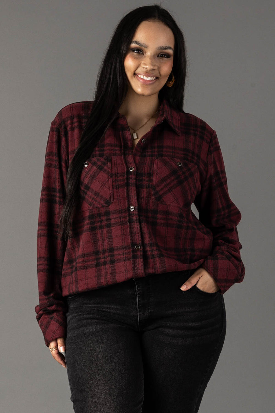 A woman with long dark hair smiles, wearing the Panache Apparel Collection Maroon & Black Plaid Button Up and black jeans, standing against a plain gray background.