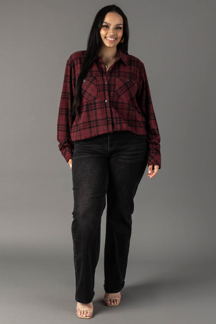A woman with long dark hair stands smiling against a plain gray background, wearing the Panache Apparel Collection’s Maroon & Black Plaid Button Up shirt, black jeans, and open-toed shoes.