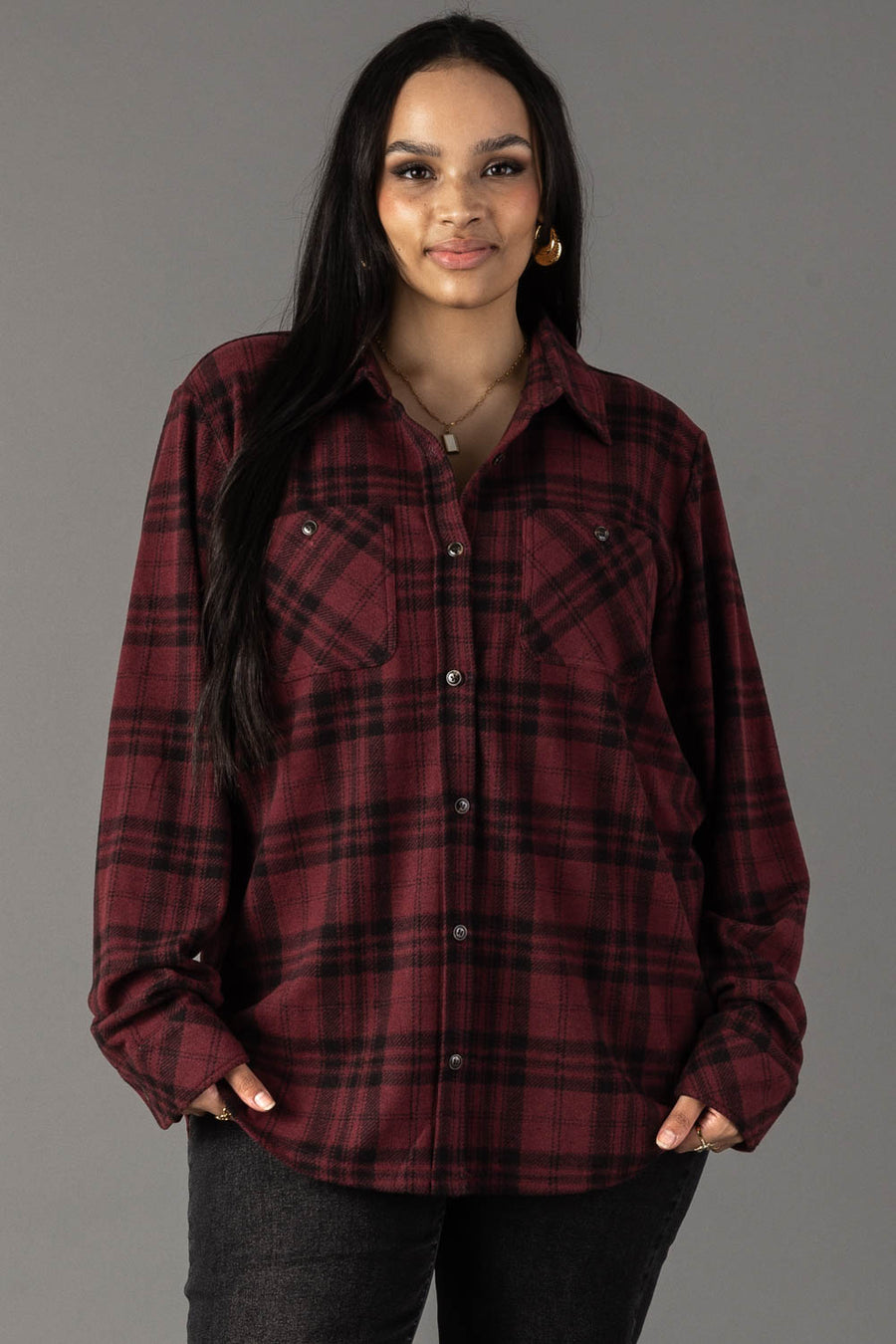 A woman wearing the Panache Apparel Collection Maroon & Black Plaid Button Up stands against a gray background, looking at the camera and smiling softly.