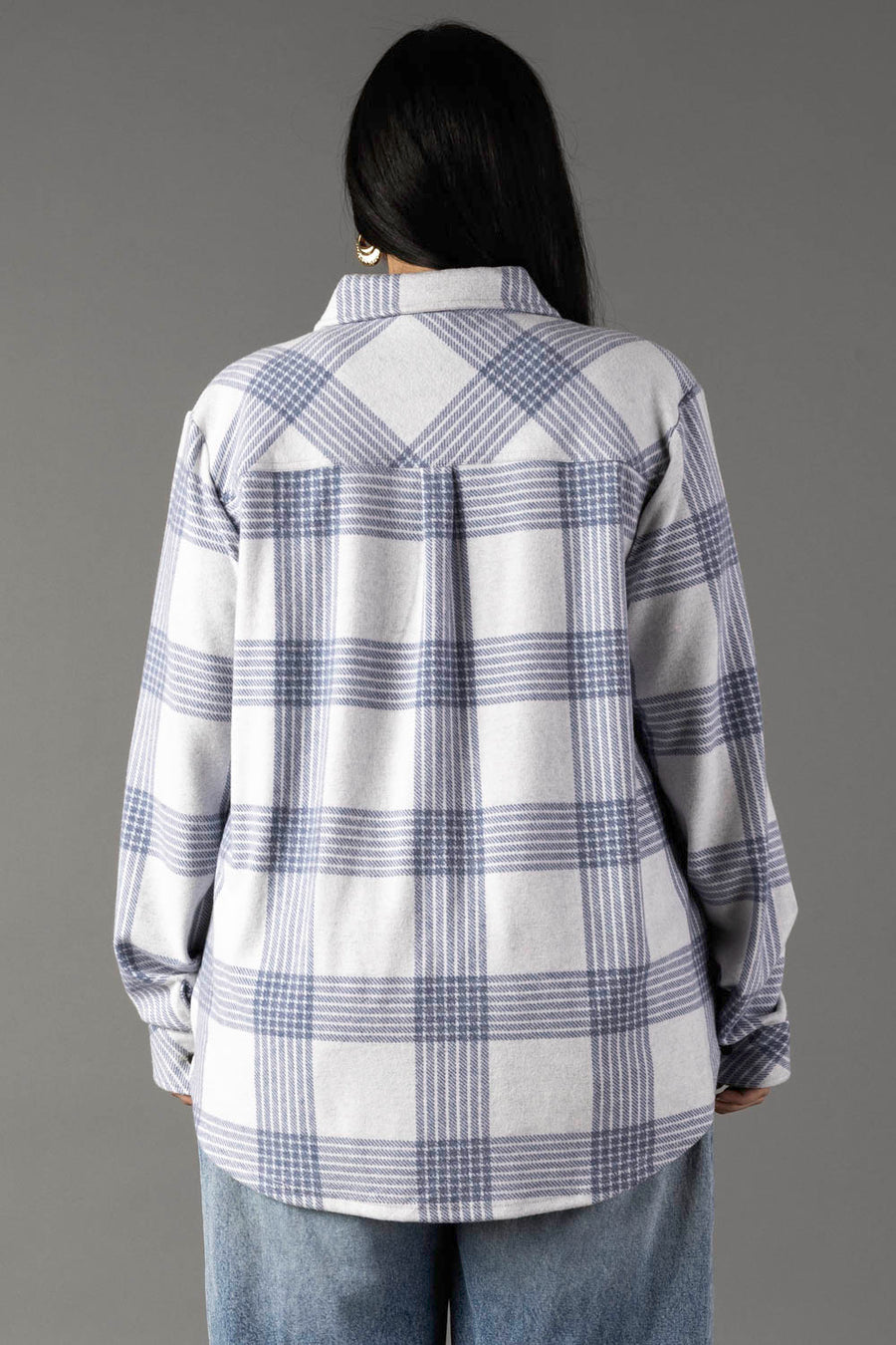 A person with long dark hair, wearing the Panache Apparel Collection’s White & Dusty Blue Plaid Button Up and light blue jeans, is standing with their back to the camera against a plain gray background.