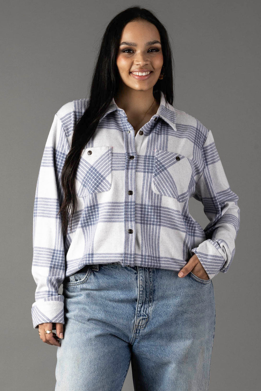 A woman with long dark hair smiles while wearing the Panache Apparel Collection White & Dusty Blue Plaid Button Up and blue jeans, standing against a plain gray background.