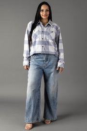 A woman with long dark hair stands against a gray background, wearing the Panache Apparel Collection’s White & Dusty Blue Plaid Button Up paired with wide-leg blue jeans and open-toed shoes.