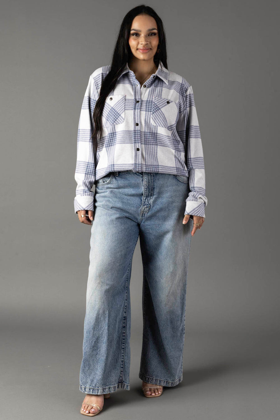 A woman with long dark hair stands against a gray background, wearing the Panache Apparel Collection’s White & Dusty Blue Plaid Button Up paired with wide-leg blue jeans and open-toed shoes.