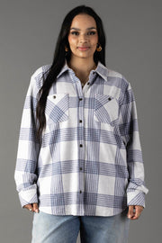 A woman with long dark hair wears the Panache Apparel Collection White & Dusty Blue Plaid Button Up, paired with light blue jeans. She stands against a plain gray background, smiling slightly at the camera.