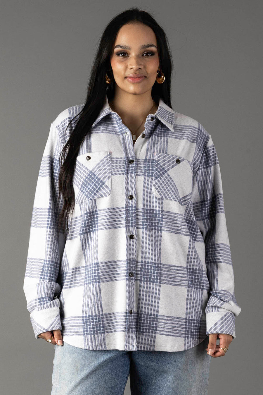 A woman with long dark hair wears the Panache Apparel Collection White & Dusty Blue Plaid Button Up, paired with light blue jeans. She stands against a plain gray background, smiling slightly at the camera.