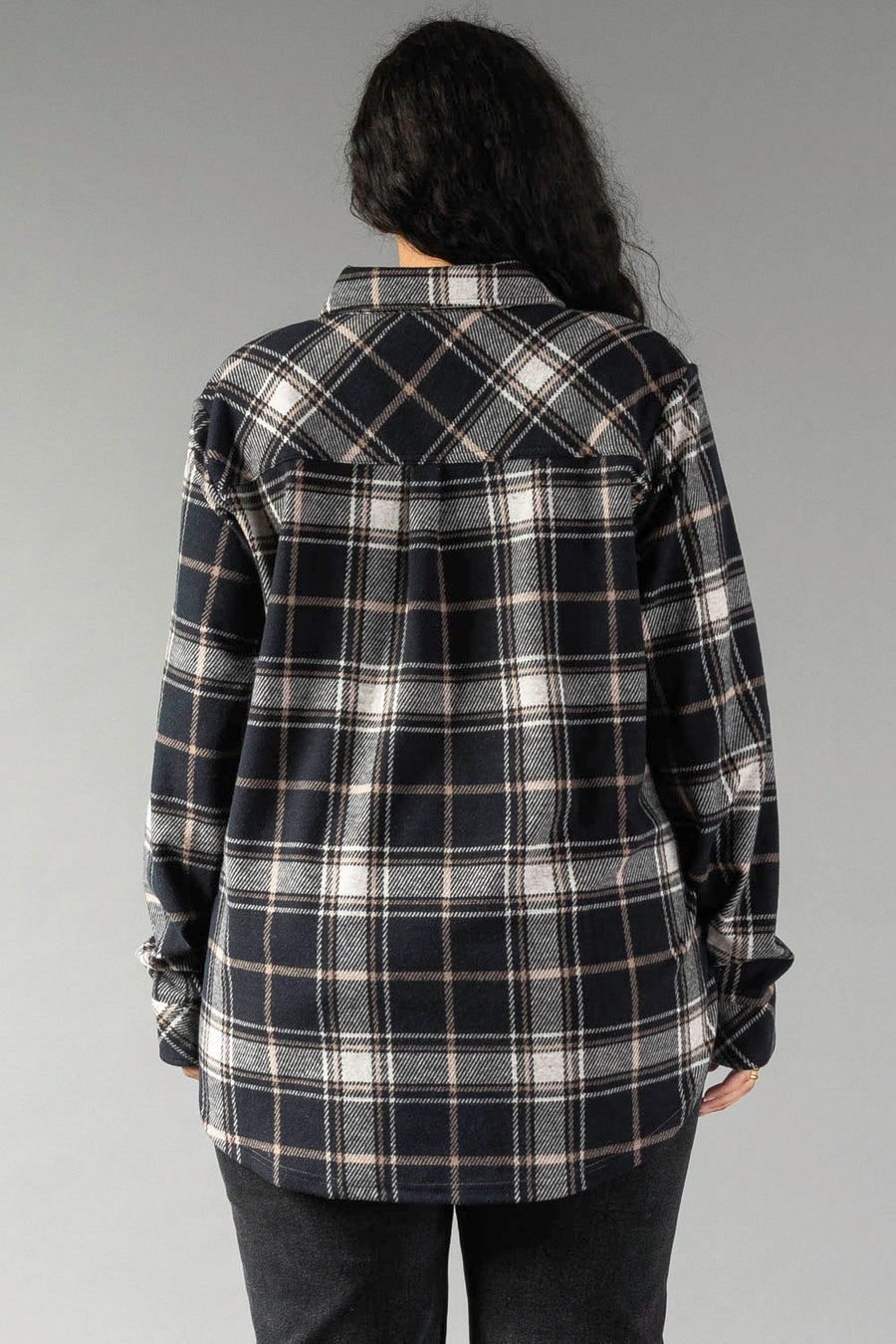 A person with long dark hair, seen from behind, wears the Panache Apparel Collection Black, White & Tan Plaid Button Up and dark pants, standing against a plain gray background.