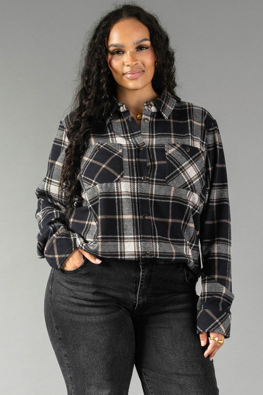 A woman with long curly hair wears the Panache Apparel Collection Black, White & Tan Plaid Button Up paired with black jeans, standing with one hand in her pocket and smiling slightly against a plain gray background.