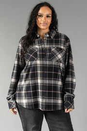 A woman with long curly hair wears the Panache Apparel Collection Black, White & Tan Plaid Button Up and black jeans, standing against a plain gray background and smiling slightly.