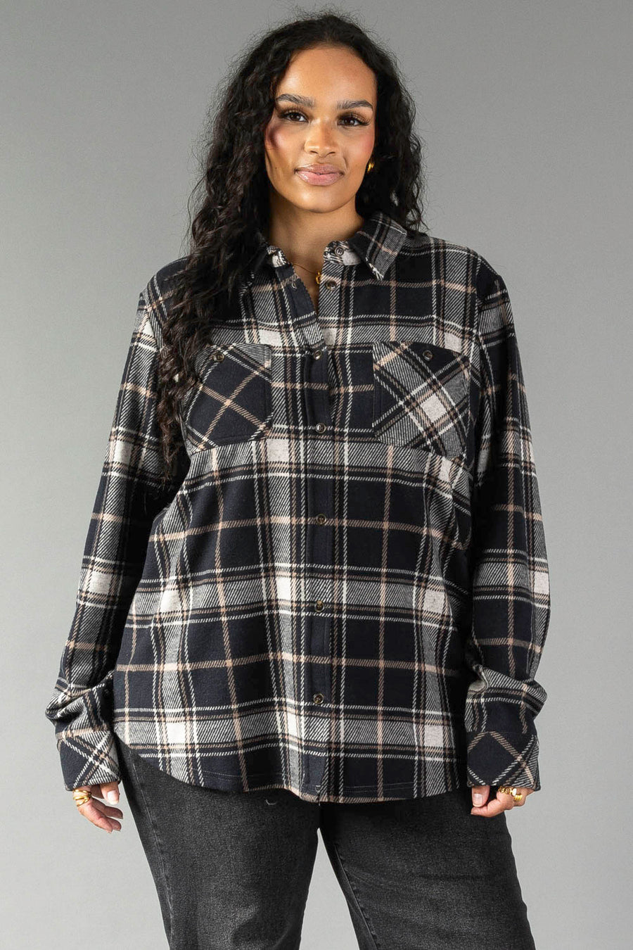 A woman with long curly hair wears the Panache Apparel Collection Black, White & Tan Plaid Button Up and black jeans, standing against a plain gray background and smiling slightly.