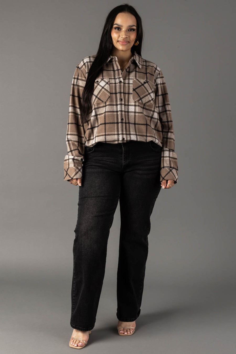 A woman stands facing forward, wearing the Panache Apparel Collection’s Tan, White & Black Plaid Button Up with black jeans and clear-heeled shoes. The background is plain gray.