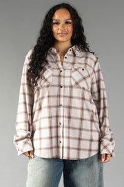 A woman with long curly hair wears the Panache Apparel Collection’s Light Taupe & Brown Plaid Button Up and blue jeans, standing against a plain gray background and smiling slightly.