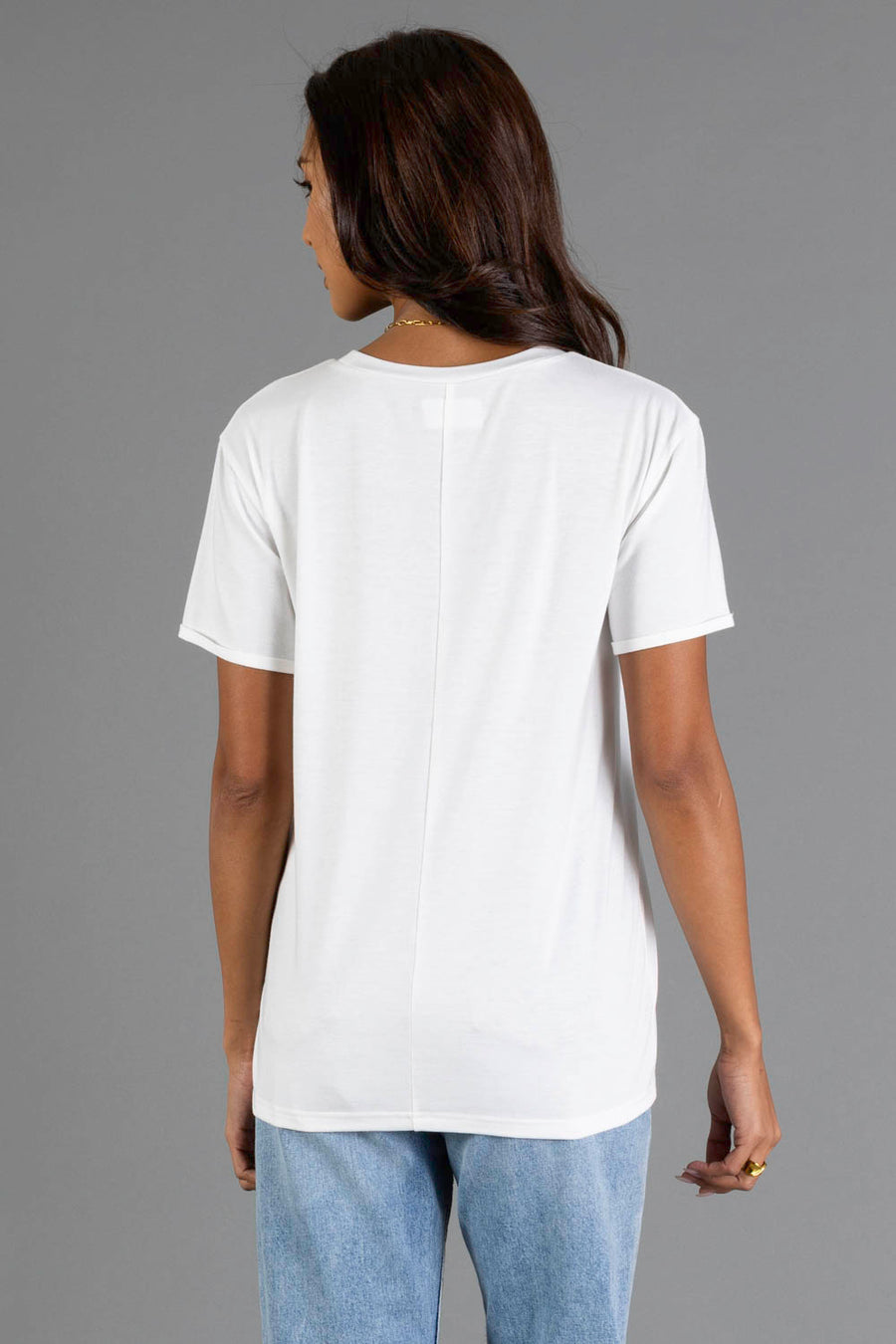 A person viewed from behind, wearing a white Merry Little Cocktail Tee and light blue jeans.