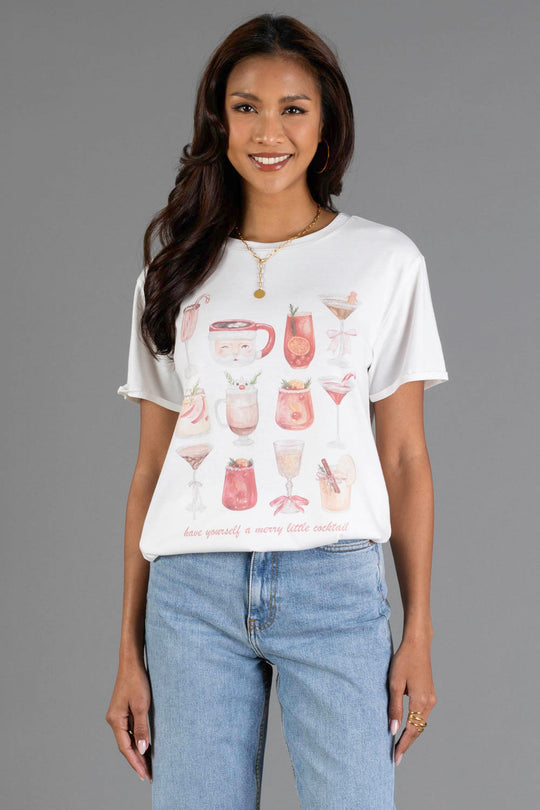 A person wears a white Merry Little Cocktail Tee featuring illustrations of holiday drinks and the text, Have yourself a merry little cocktail.