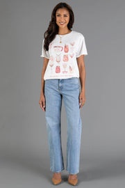 A person wearing a white Merry Little Cocktail Tee featuring holiday illustrations and the text have yourself a merry little cocktail, paired with light-wash blue jeans.