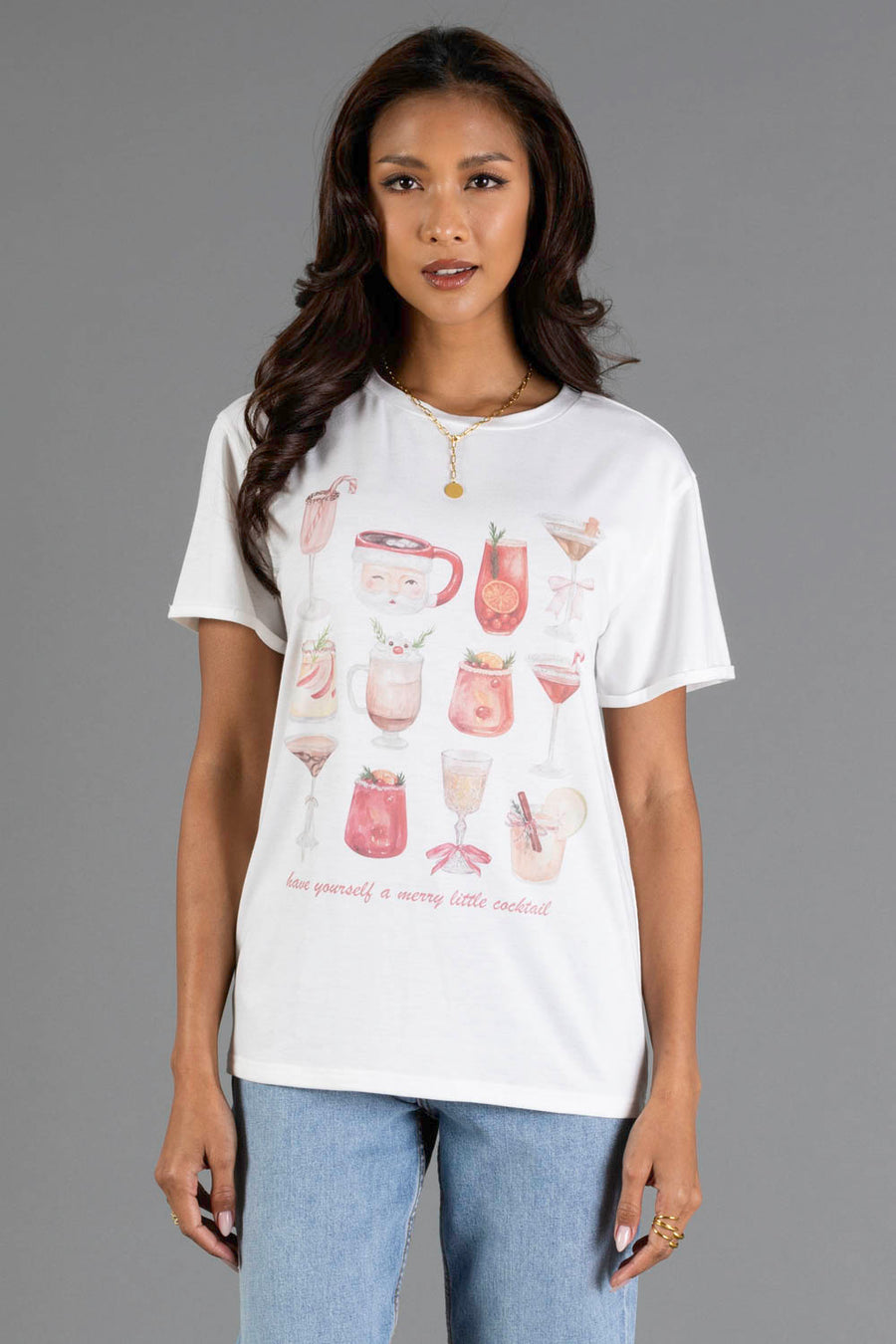 A woman wears a white Merry Little Cocktail Tee featuring a festive watercolor print of various drinks and the text have yourself a merry little cocktail.