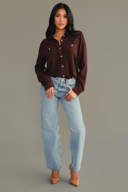 A woman stands against a gray background, wearing the Espresso Brown Super Soft Button Up from Panache Apparel Collection, paired with light blue jeans and tan pointed-toe heels. She poses with one hand in her pocket and the other resting by her side.