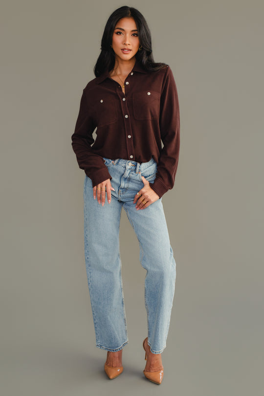 A woman stands against a gray background, wearing the Espresso Brown Super Soft Button Up from Panache Apparel Collection, paired with light blue jeans and tan pointed-toe heels. She poses with one hand in her pocket and the other resting by her side.