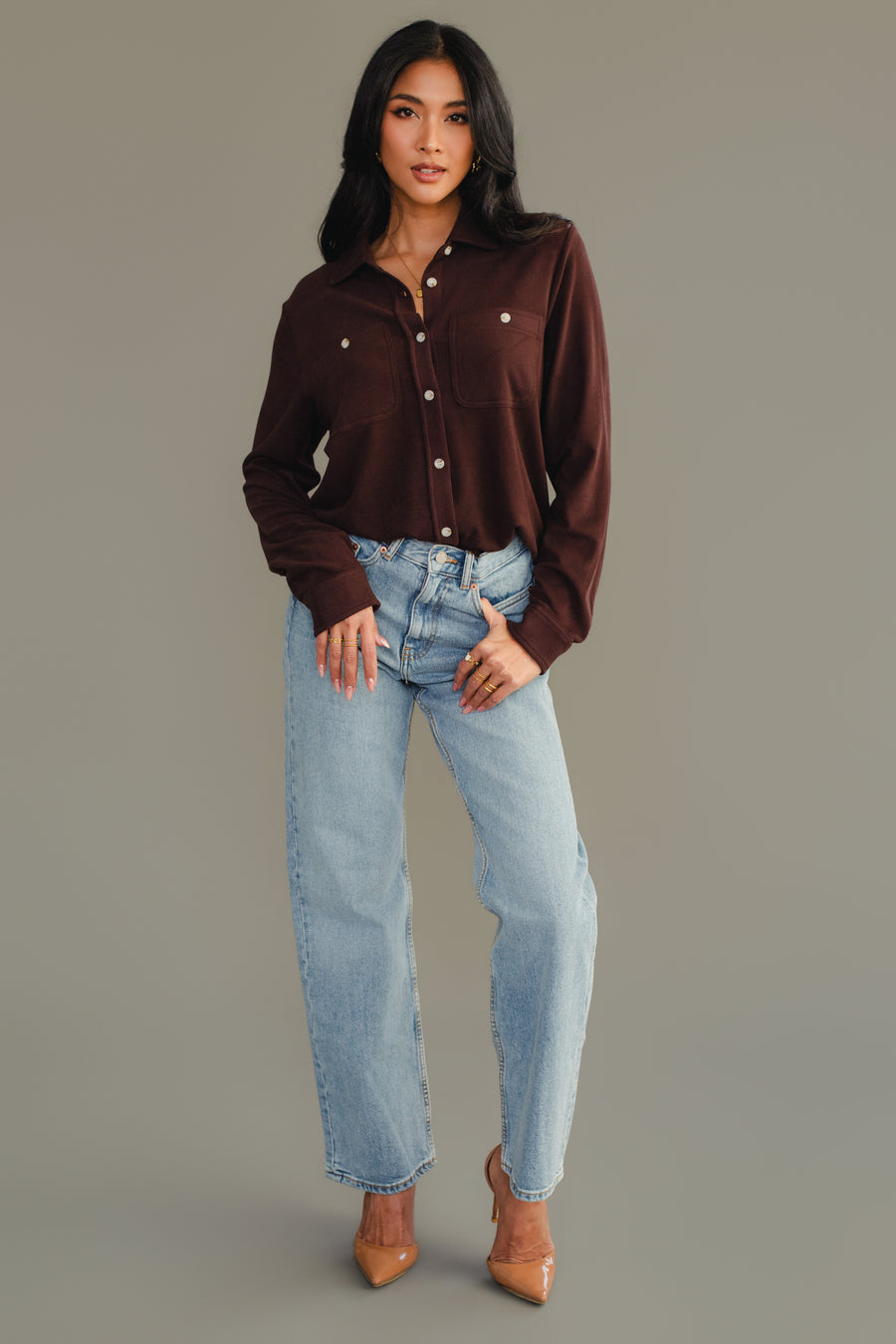 A woman stands against a gray background, wearing the Espresso Brown Super Soft Button Up from Panache Apparel Collection, paired with light blue jeans and tan pointed-toe heels. She poses with one hand in her pocket and the other resting by her side.