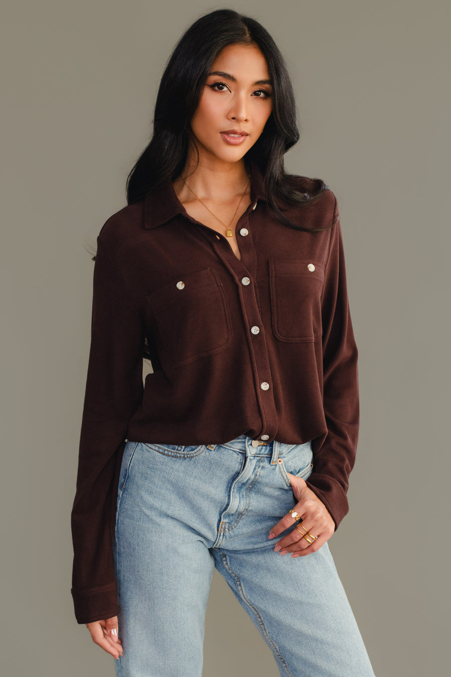 A woman with long dark hair wears the Panache Apparel Collection Espresso Brown Super Soft Button Up with chest pockets and light blue jeans, standing against a plain gray background.