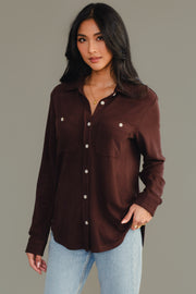 A woman models the Panache Apparel Collection’s Espresso Brown Super Soft Button Up, featuring front pockets, paired with light blue jeans. She stands against a plain gray background and looks at the camera with a slight smile.