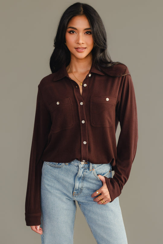 A woman with long dark hair wears the Espresso Brown Super Soft Button Up from Panache Apparel Collection and light blue jeans, standing against a plain gray background and looking at the camera with a relaxed expression.